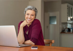 Beautiful senior woman using laptop at home. Mature smiling woman looking at camera while working with computer. Portrait of happy modern retired lady with laptop sitting on living room.