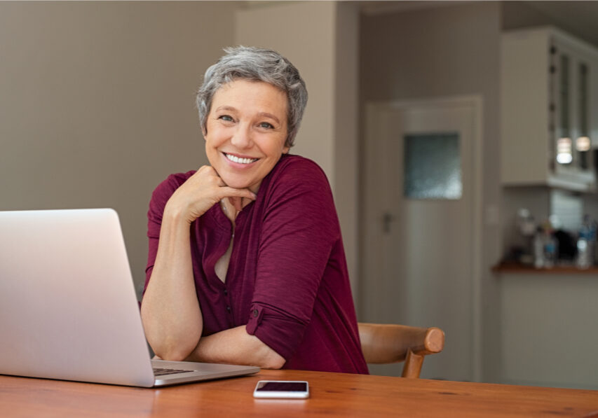 Beautiful senior woman using laptop at home. Mature smiling woman looking at camera while working with computer. Portrait of happy modern retired lady with laptop sitting on living room.